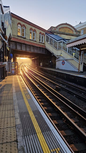 The image depicts a railway station platform at sunset, with the train tracks running into a tunnel illuminated by the warm glow of the setting sun. The platform surface features tactile paving strips